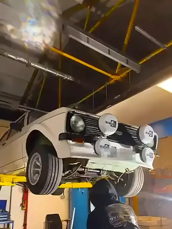 A white classic Ford raised on a two-post ramp in a workshop, with an operator in a dark hoodie dry ice blasting underneath it.