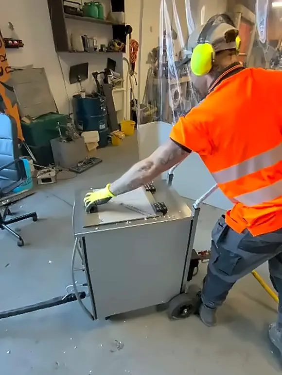 An operator in an orange hi-vis vest, yellow ear defenders and gloves closing the hopper lid on a dry ice blaster in a workshop.